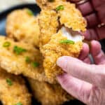 Hands pulling apart an air fryer chicken tender that's juicy on the inside and crispy on the outside, with a plate of chicken in the background.
