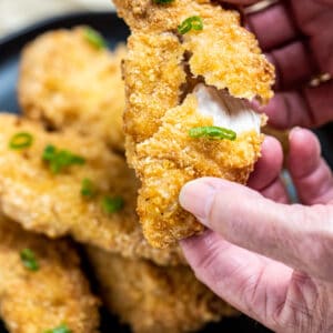 Hands pulling apart an air fryer chicken tender that's juicy on the inside and crispy on the outside, with a plate of chicken in the background.