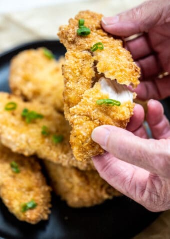 Hands pulling apart an air fryer chicken tender that's juicy on the inside and crispy on the outside, with a plate of chicken in the background.
