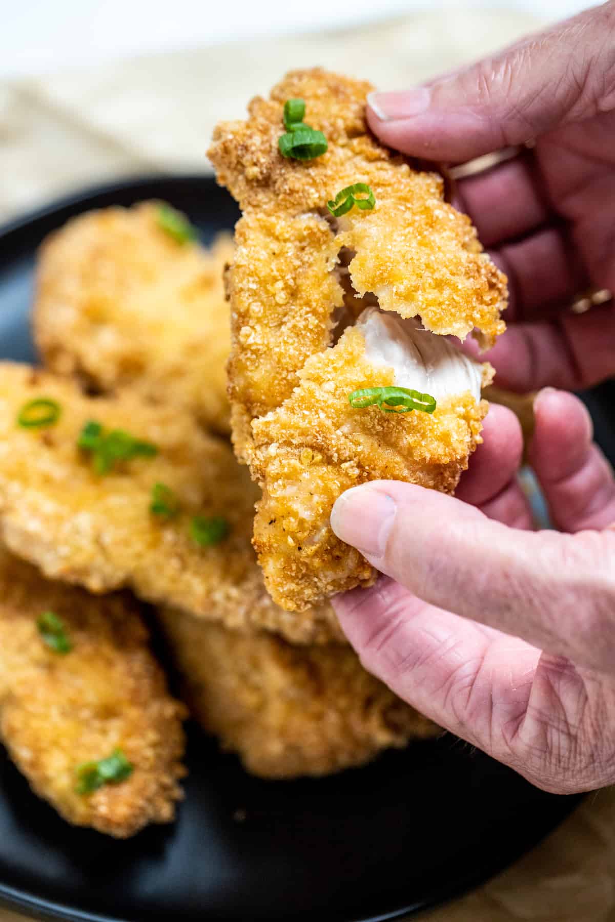 Hands pulling apart an air fryer chicken tender that's juicy on the inside and crispy on the outside, with a plate of chicken in the background.