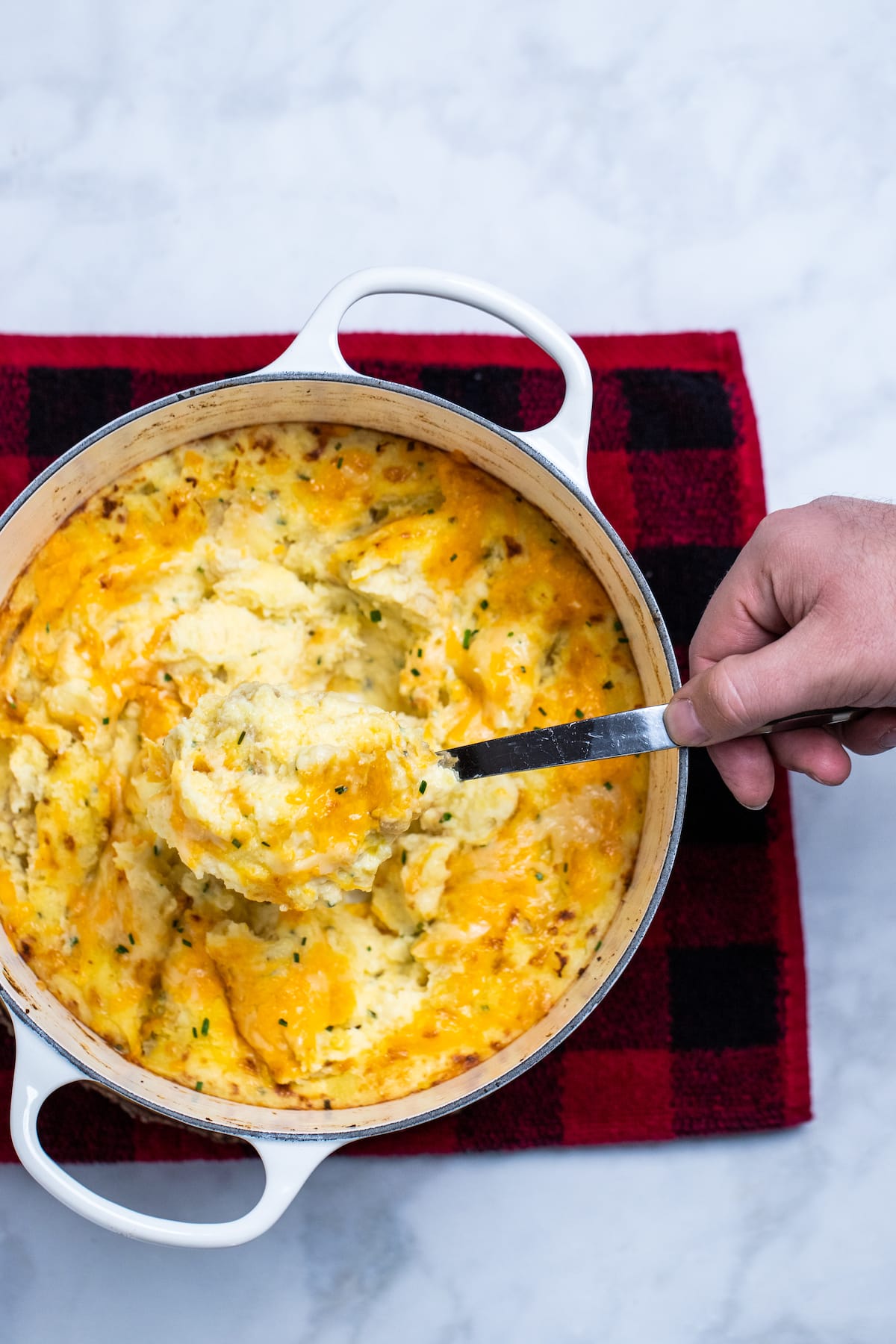 A dutch oven with cheesy mashed potatoes on top of a plaid towel, with a hand using a serving spoon to scoop out potatoes.