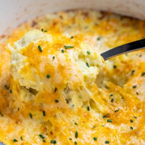 A dutch oven with a spoon scooping cheesy mashed potatoes, with melty cheese pulling from the dish.
