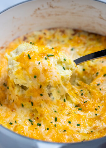 A dutch oven with a spoon scooping cheesy mashed potatoes, with melty cheese pulling from the dish.