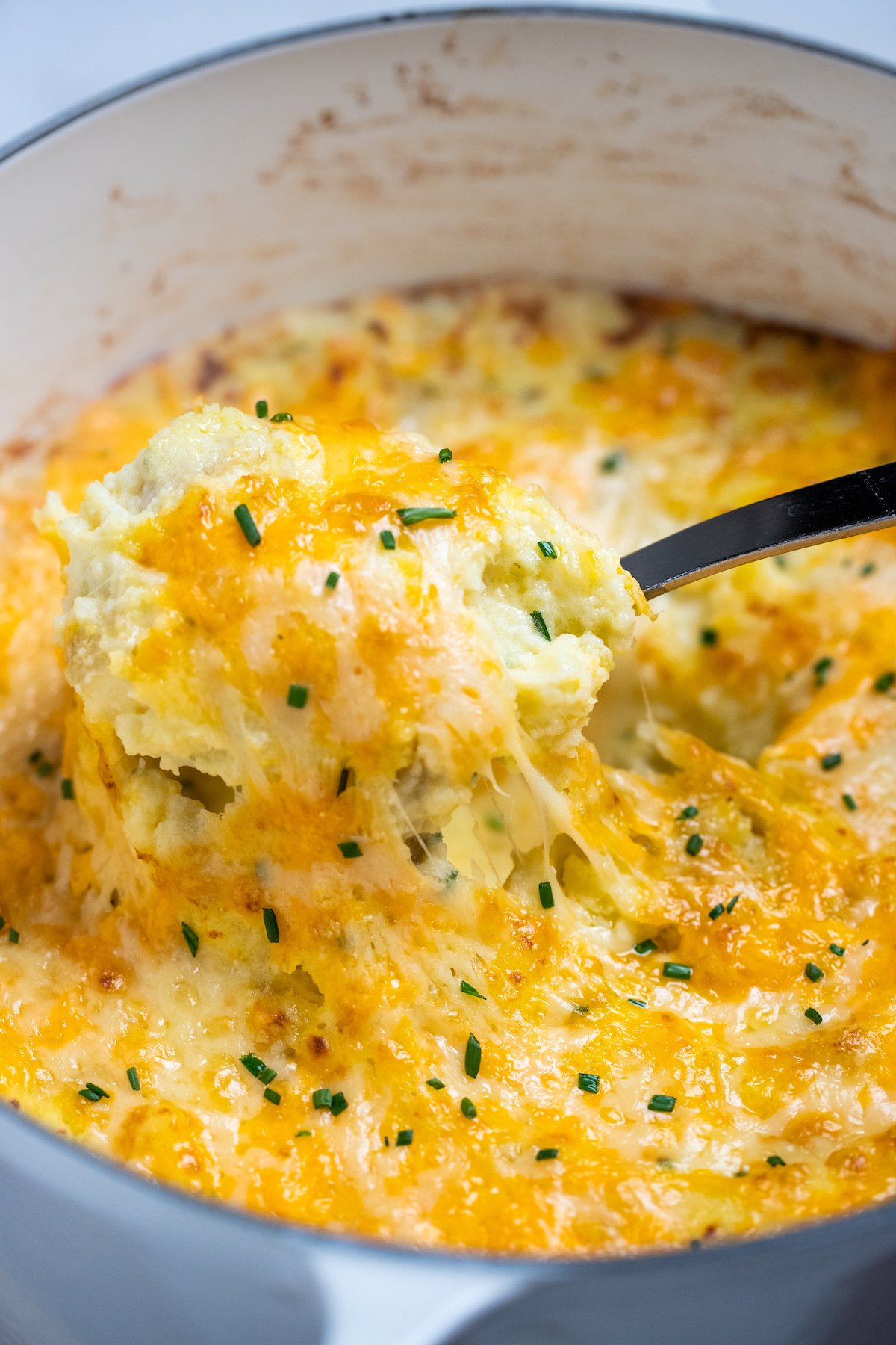 A dutch oven with a spoon scooping cheesy mashed potatoes, with melty cheese pulling from the dish.