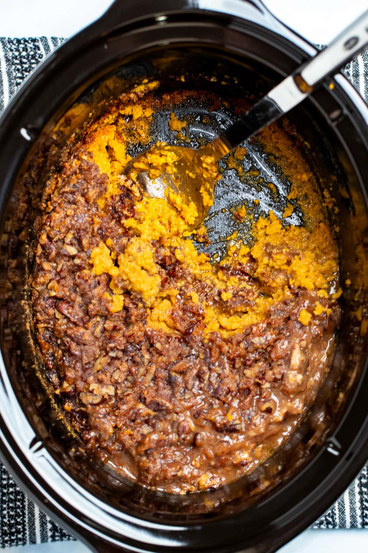 A crockpot sitting on top of a towel on the table, with a spoon sitting in the pot having already served some of the sweet potato casserole.