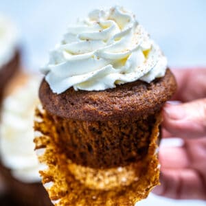 A hand holding a gingerbread cupcake with frosting, with the paper liner half peeled off.