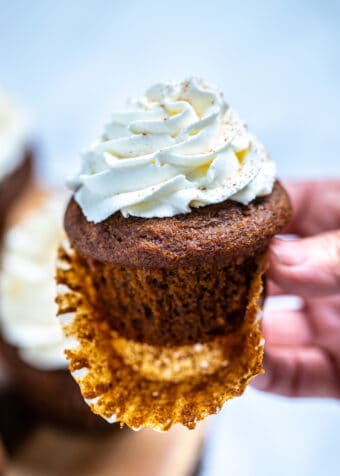 A hand holding a gingerbread cupcake with frosting, with the paper liner half peeled off.