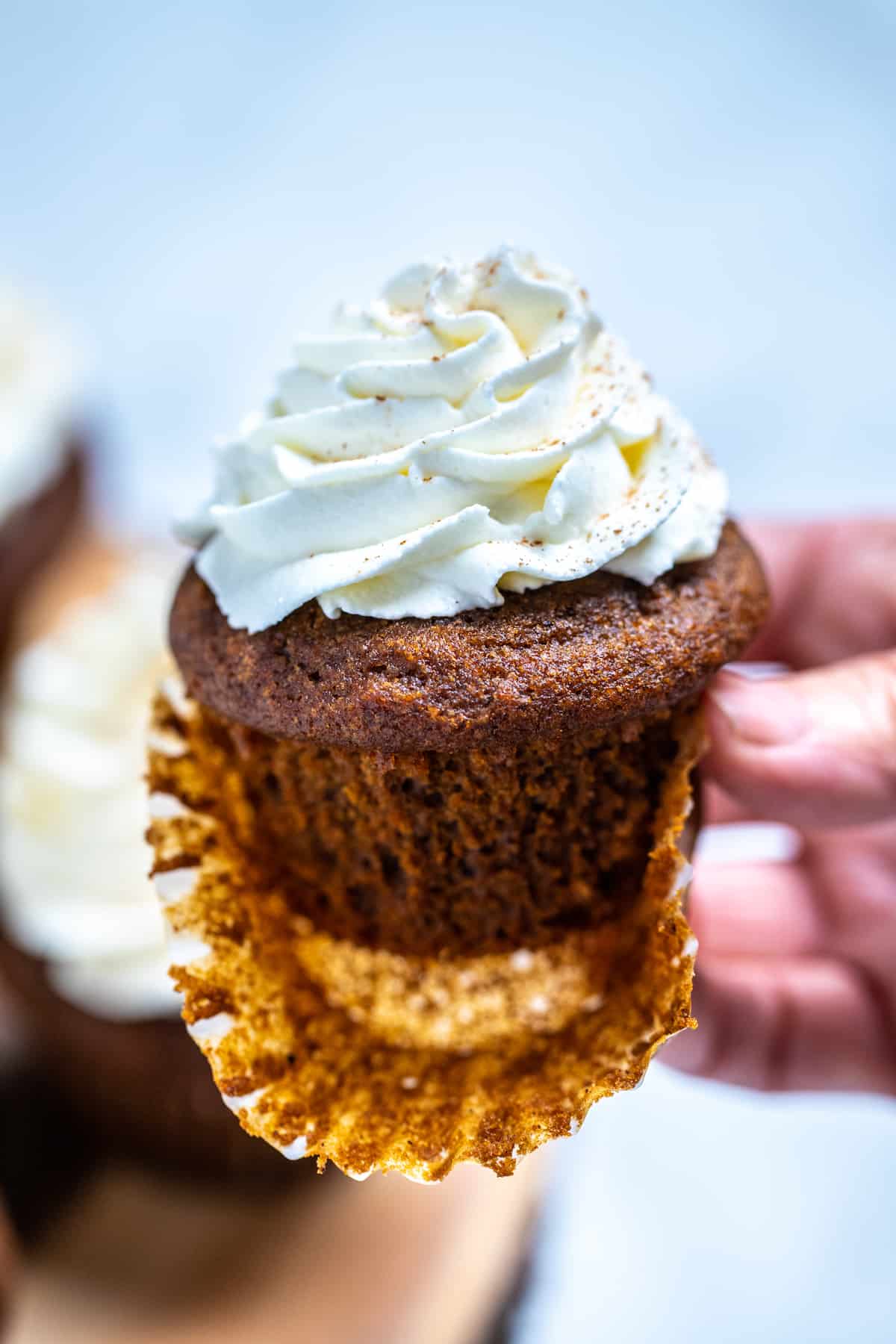 A hand holding a gingerbread cupcake with frosting, with the paper liner half peeled off.