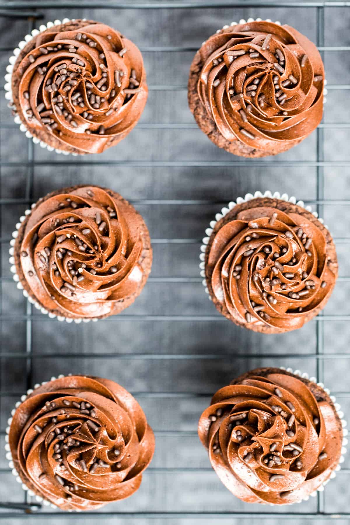 Chocolate cupcakes with frosting topped with chocolate sprinkles on top of a wire rack.