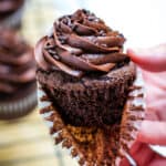 Two hands pulling the paper liner off of a gluten free chocolate cupcake with more cupcakes in the background.