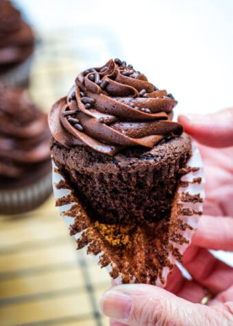 Two hands pulling the paper liner off of a gluten free chocolate cupcake with more cupcakes in the background.