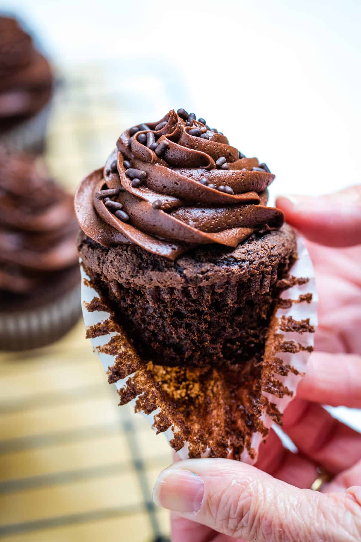 Two hands pulling the paper liner off of a gluten free chocolate cupcake with more cupcakes in the background.