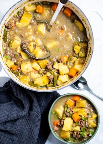 A bowl of ground beef soup next to a spoon and a kitchen towel, in front of a dutch oven full of soup with a ladle in it.