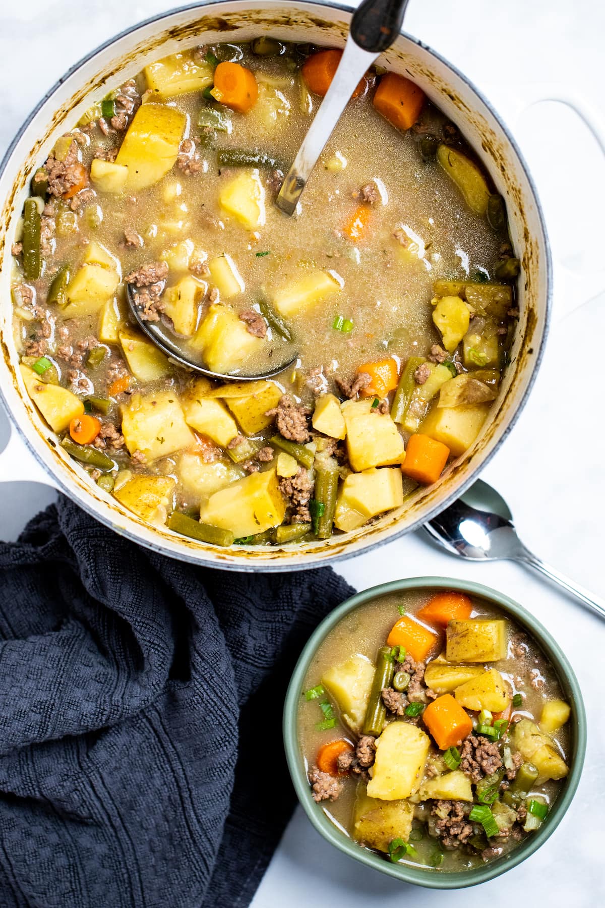 A bowl of ground beef soup next to a spoon and a kitchen towel, in front of a dutch oven full of soup with a ladle in it.