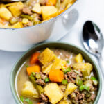 A bowl of ground beef soup in front of a dutch oven of soup with a spoon next to the bowl.