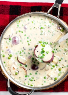 A skillet with creamed peas and potatoes on top of a plaid towel, with a ladle scooping some up.