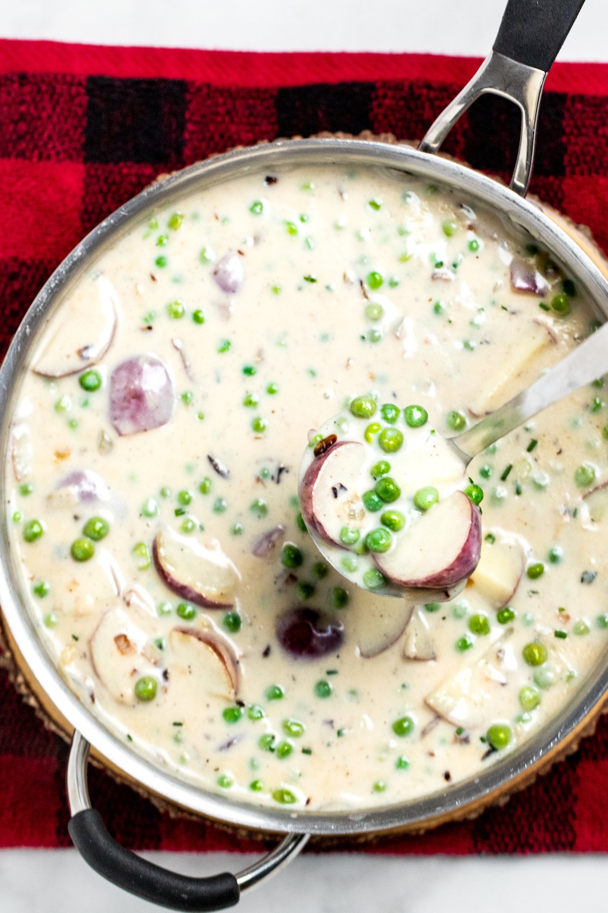 A skillet with creamed peas and potatoes on top of a plaid towel, with a ladle scooping some up.