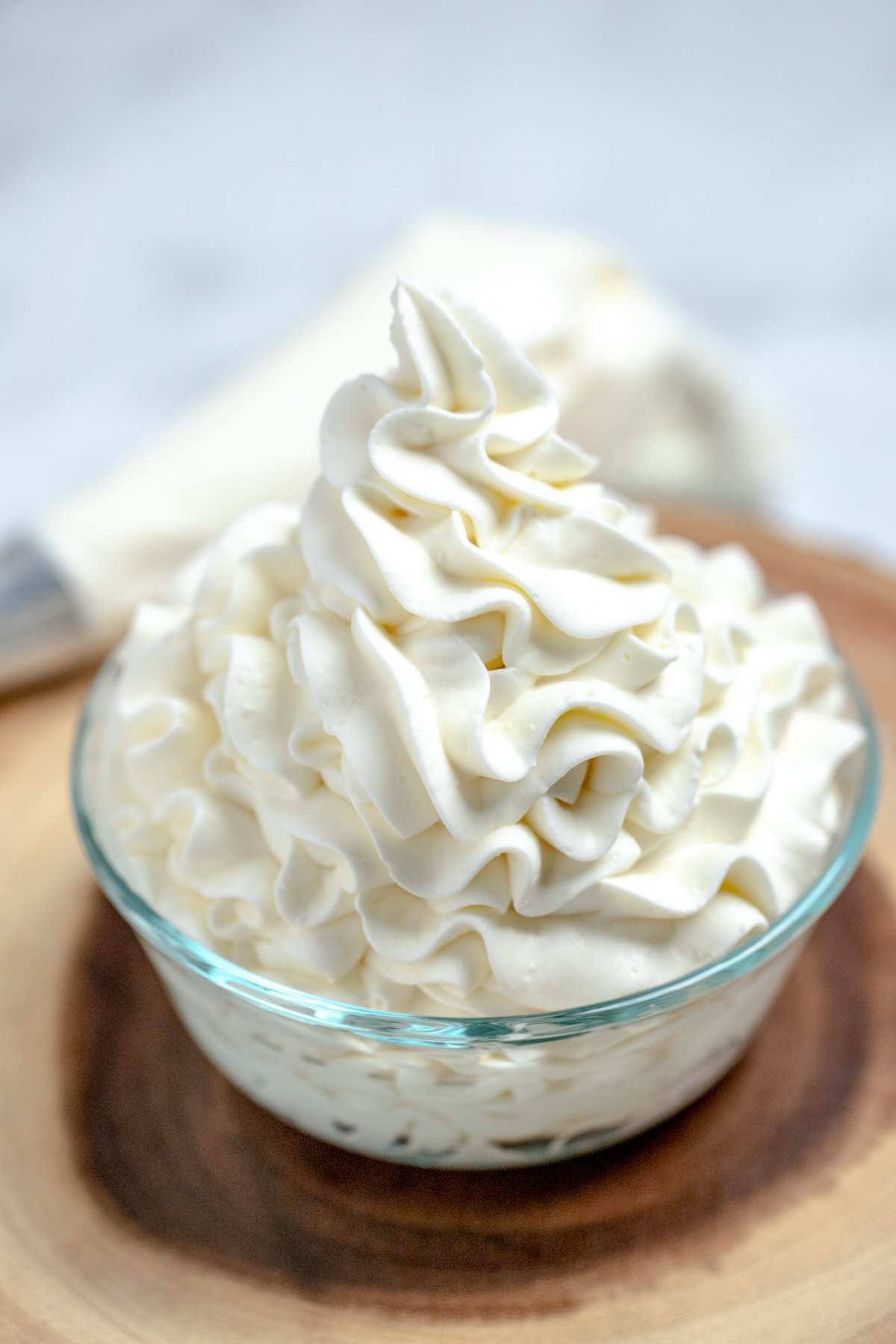 Whipped cream cheese frosting piped into a glass bowl on a wooden cutting board with a piping bag in the background.