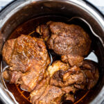 An instant pot with cooked pork shoulder on a trivet with drippings below.