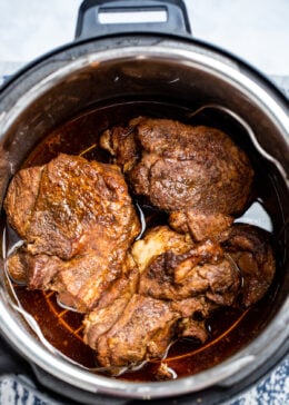 An instant pot with cooked pork shoulder on a trivet with drippings below.