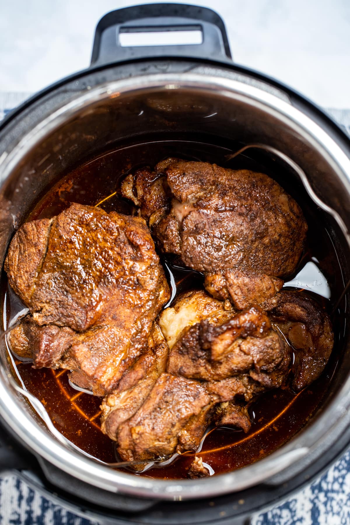 An instant pot with cooked pork shoulder on a trivet with drippings below.