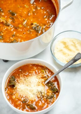 A bowl of kale and white bean soup topped with parmesan cheese, with a spoon resting in it, next to a dish of parmesan cheese and in front of a large pot of soup.