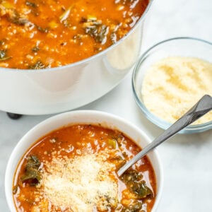 A bowl of kale and white bean soup topped with parmesan cheese, with a spoon resting in it, next to a dish of parmesan cheese and in front of a large pot of soup.