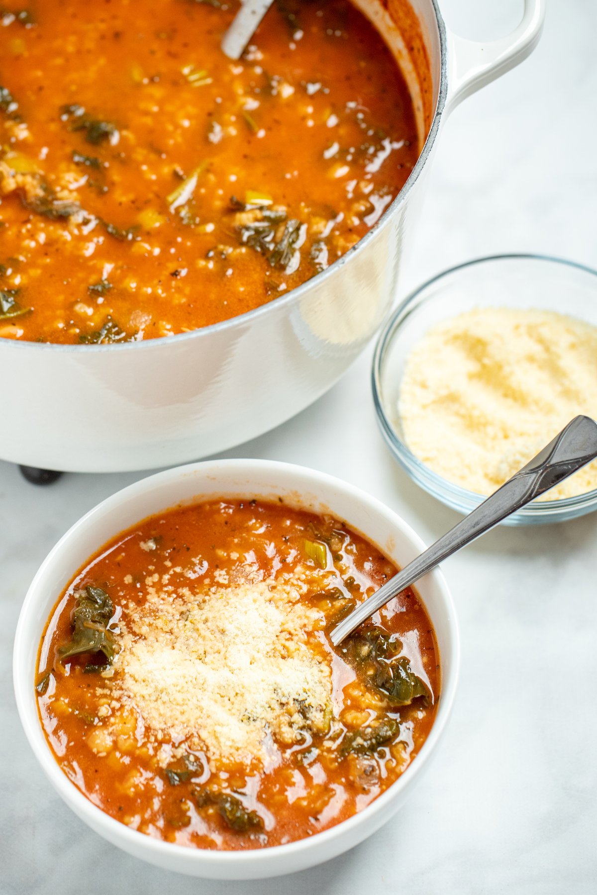 A bowl of kale and white bean soup topped with parmesan cheese, with a spoon resting in it, next to a dish of parmesan cheese and in front of a large pot of soup.