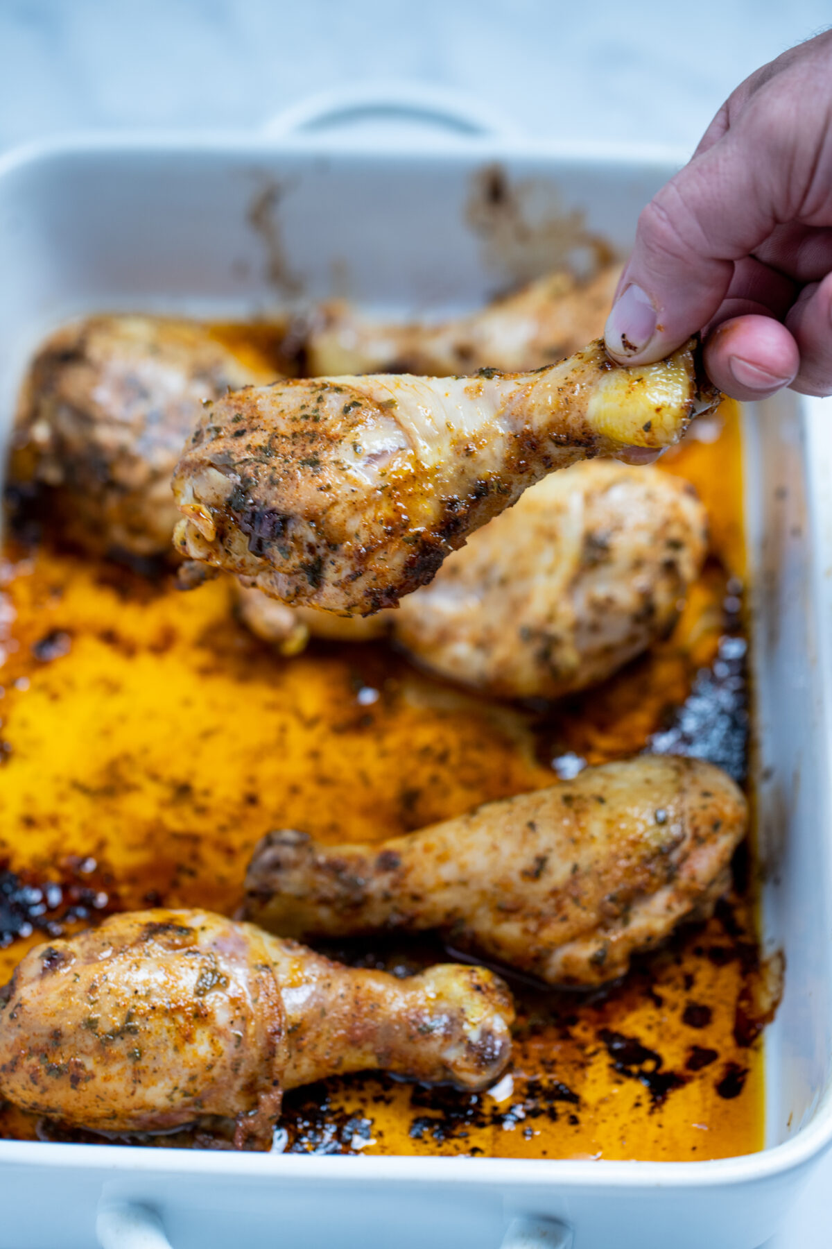 A baking dish with cooked chicken drumsticks covered in marinade with a hand picking up one drumstick.