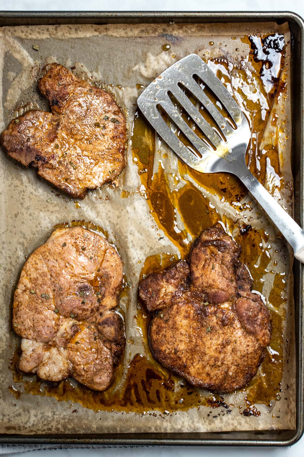 A sheet pan lined with parchment paper with marinated pork chops fully cooked, and a spatula in the upper right hand corner.