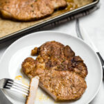 A marinated pork chop on a plate with a fork holding a slice of chop, next to a sheet pan with other cooked pork chops.