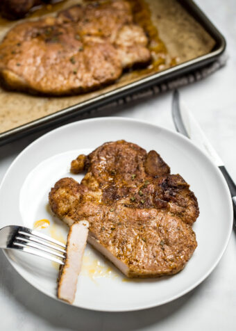A marinated pork chop on a plate with a fork holding a slice of chop, next to a sheet pan with other cooked pork chops.