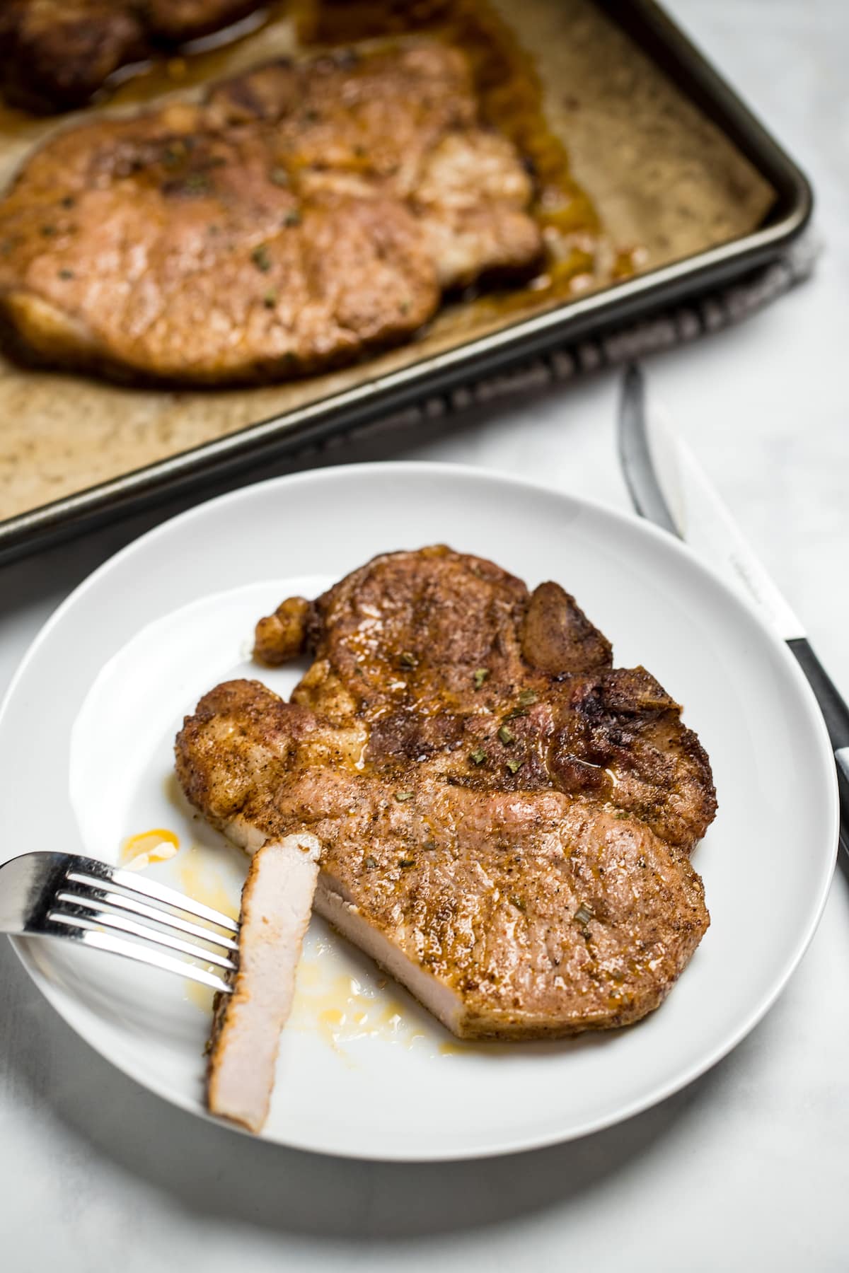 A marinated pork chop on a plate with a fork holding a slice of chop, next to a sheet pan with other cooked pork chops.