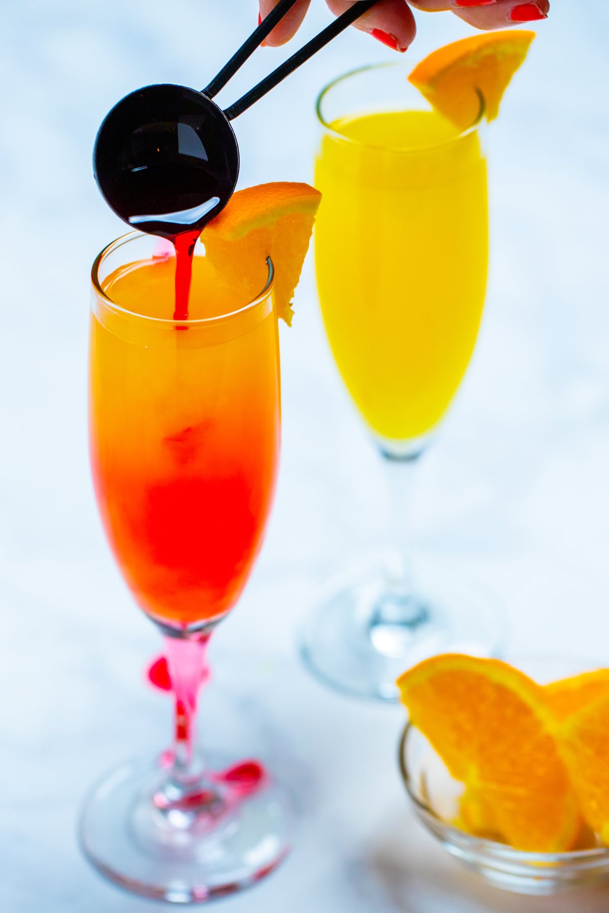 Two wine flutes with prosecco mimosas garnished with orange slices, and a tablespoon pouring grenadine into one of the glasses, with a small glass bowl of oranges on the table next to them.