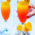 Two wine flutes with prosecco mimosas garnished with orange slices, one being held by a hand, with a small glass bowl of oranges on the table next to them.