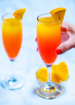 Two wine flutes with prosecco mimosas garnished with orange slices, one being held by a hand, with a small glass bowl of oranges on the table next to them.