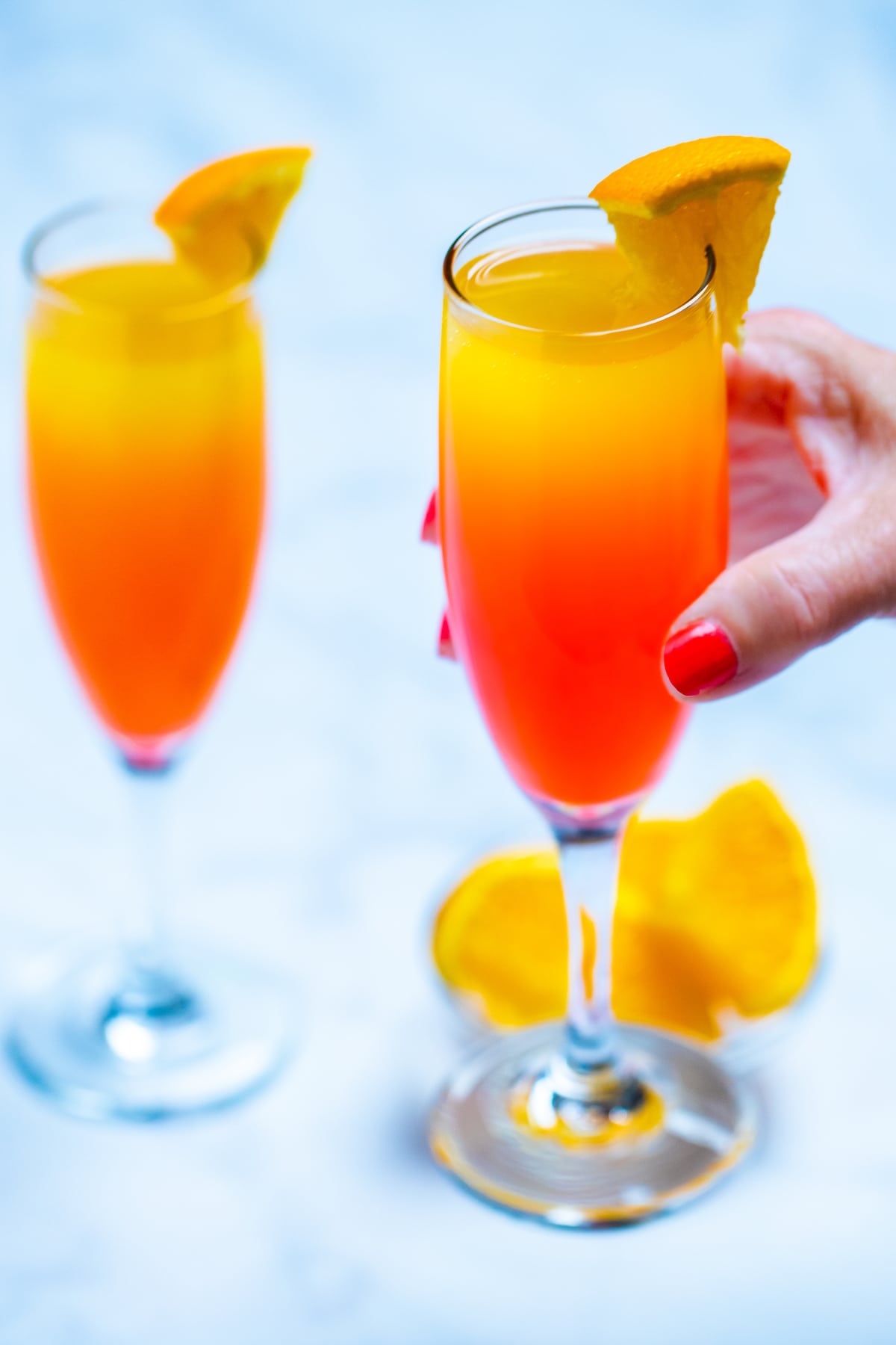Two wine flutes with prosecco mimosas garnished with orange slices, one being held by a hand, with a small glass bowl of oranges on the table next to them.