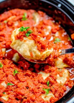 A spoon lifting a stuffed cabbage roll out of the slow cooker, topped with tomato sauce and fresh parsley.
