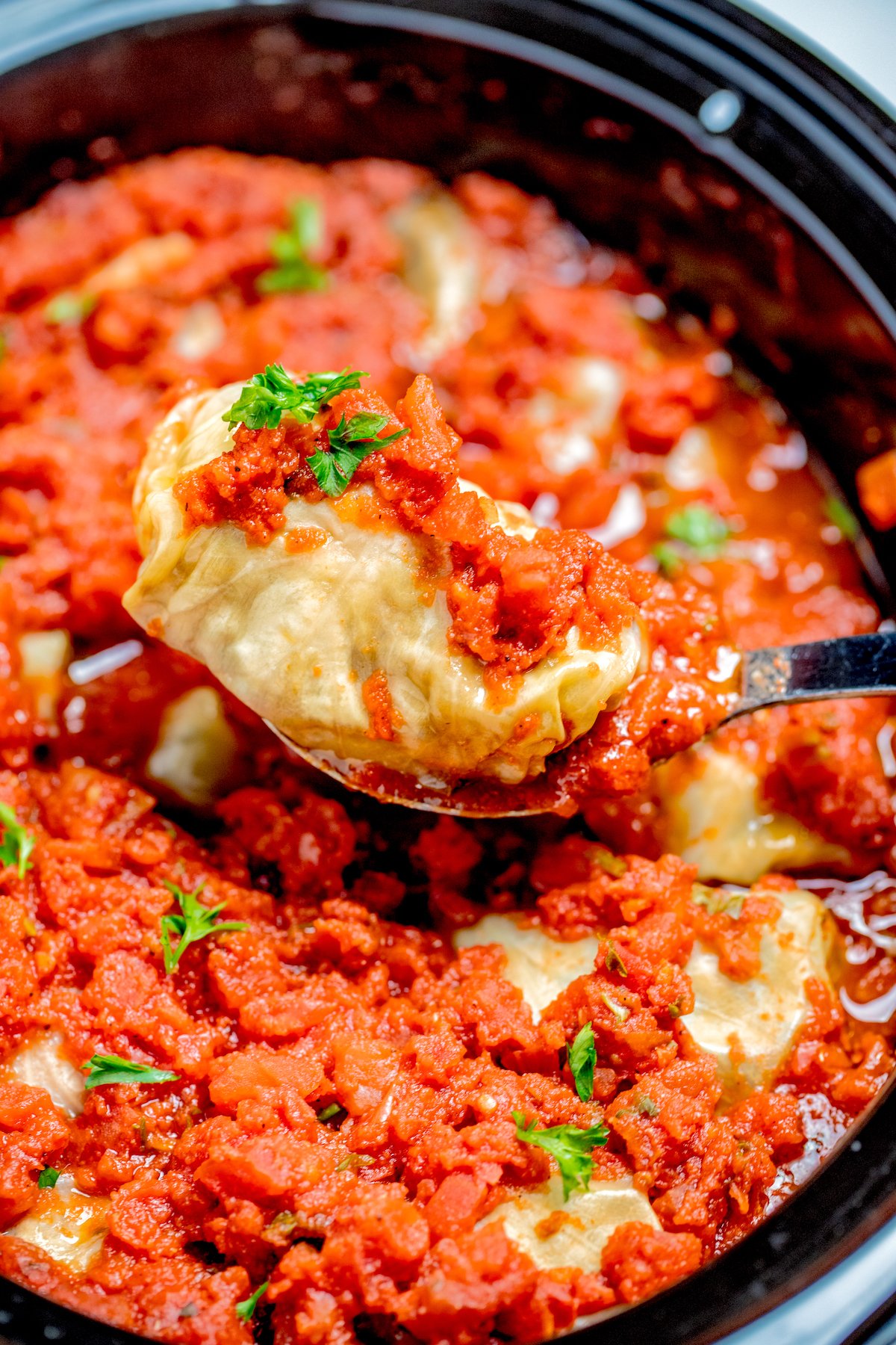 A spoon lifting a stuffed cabbage roll out of the slow cooker, topped with tomato sauce and fresh parsley.