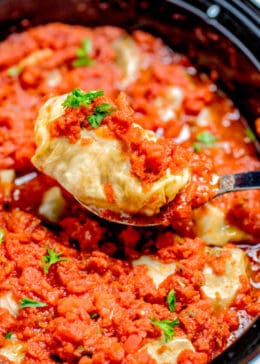 A spoon lifting a stuffed cabbage roll out of the slow cooker, topped with tomato sauce and fresh parsley.