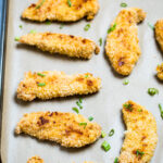 Baked chicken tenders looking golden brown and crispy on a sheet pan with parchment paper topped with green onions.