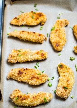 Baked chicken tenders looking golden brown and crispy on a sheet pan with parchment paper topped with green onions.