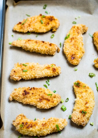 Baked chicken tenders looking golden brown and crispy on a sheet pan with parchment paper topped with green onions.