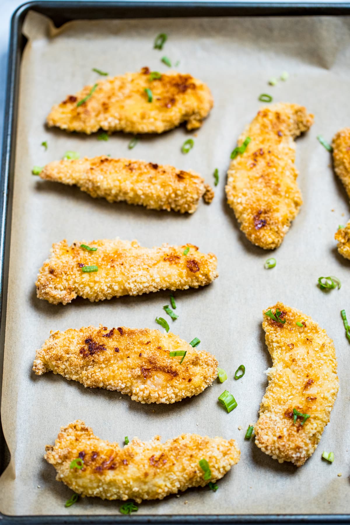 Baked chicken tenders looking golden brown and crispy on a sheet pan with parchment paper topped with green onions.