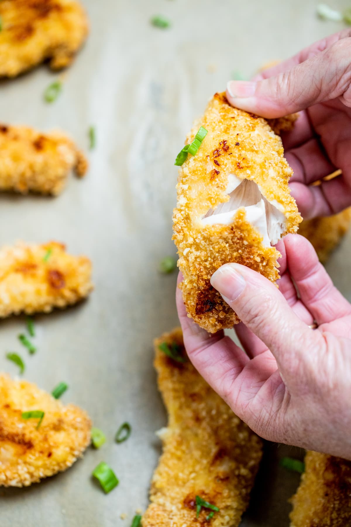 A hand pulling apart a baked chicken tender over a sheet pan of other chicken tenders topped with green onions.