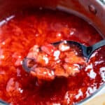 A spoon lifting strawberry cake filling out of a saucepan of filling.