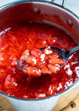 A spoon lifting strawberry cake filling out of a saucepan of filling.