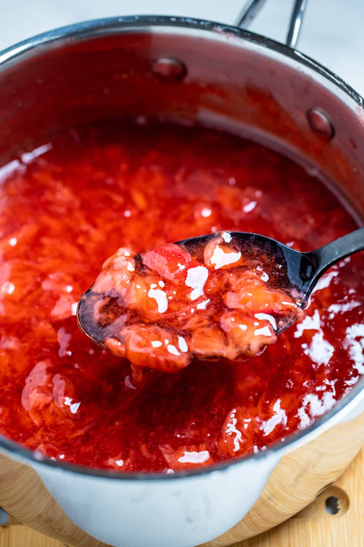 A spoon lifting strawberry cake filling out of a saucepan of filling.