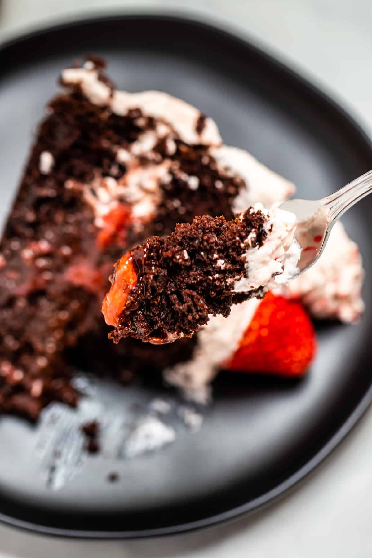 A fork full of chocolate cake with strawberry filling over a plate with a slice of cake.