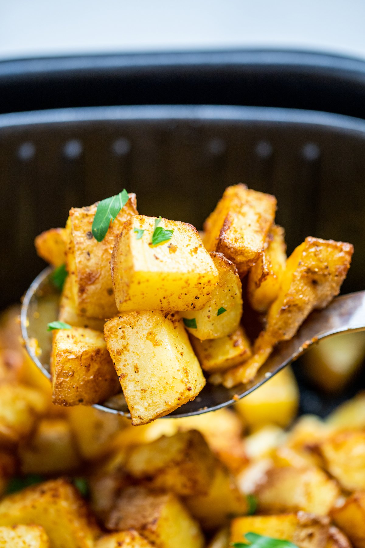 A spoon lifting breakfast potatoes out of the air fryer basket.